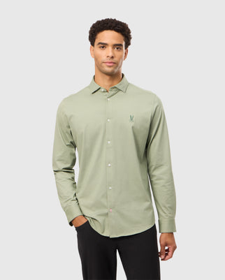 A person is wearing a MENS BRENDAN JERSEY SHIRT - B6C749D200 from Psycho Bunny. This light green button-up shirt has long sleeves and is made from soft Pima cotton jersey, featuring the brand's embroidered chest Bunny logo. They casually have one hand in their pocket while standing against a plain white background.