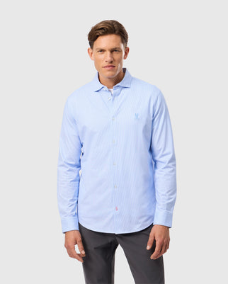 A man wearing a light blue Psycho Bunny MENS BROOK STRIPED JERSEY SHIRT - B6C750D200 and dark pants stands against a plain white background. The comfortable shirt features a small embroidered logo on the left side. He has short brown hair and a neutral expression.