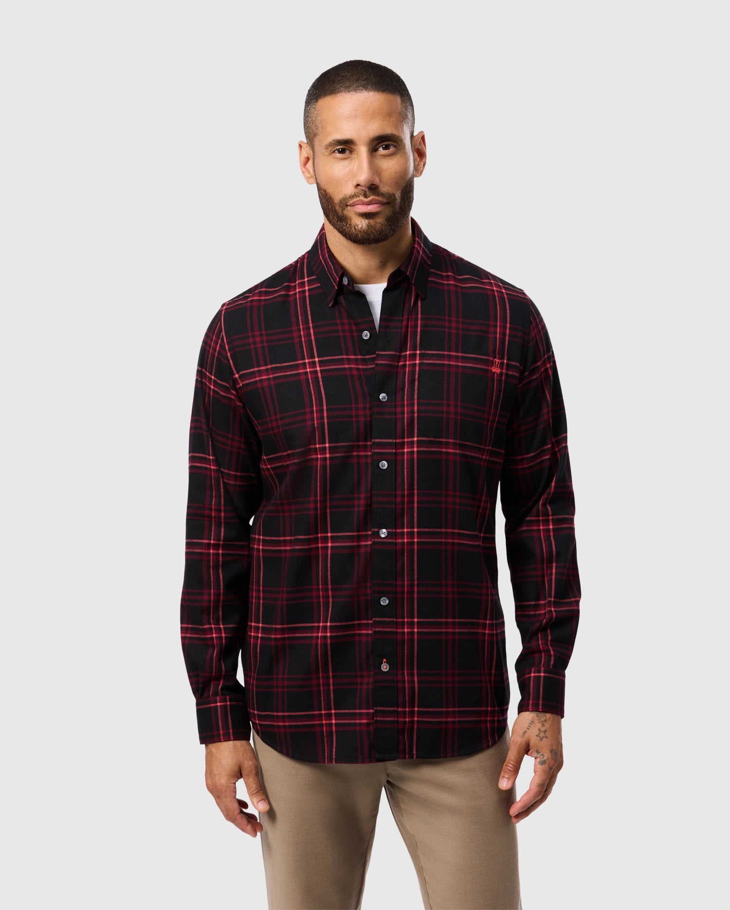 A man with short hair and a beard is wearing the Psycho Bunny MENS HAYES PLAID SHIRT - B6C842D200, which features a red and black plaid pattern made of Pima cotton. He pairs it with beige pants while standing against a plain light gray background.