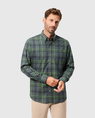 A man with short brown hair and a beard is wearing the Psycho Bunny MENS HAYES PLAID SHIRT - B6C842D200 in green and black, paired with beige pants, standing against a plain gray background. His hands are loosely clasped as he looks forward.