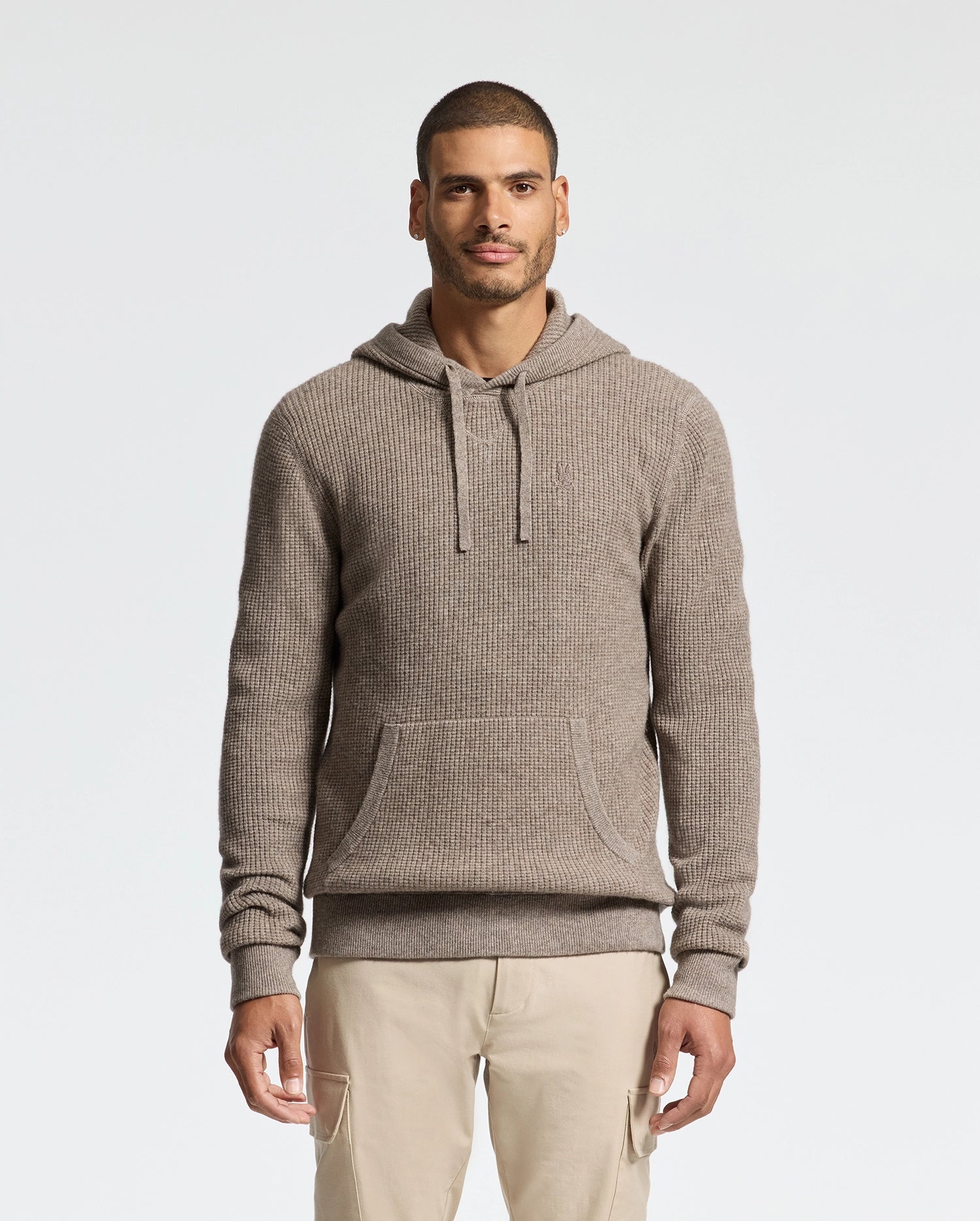 A man in a beige Psycho Bunny MENS LEVAN WAFFLE HOODED SWEATER stands against a plain white background, wearing cream pants. He has short hair and a trimmed beard, with his hands by his sides.