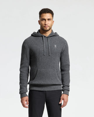 A man stands against a white background, wearing the Psycho Bunny MENS LEVAN WAFFLE HOODED SWEATER (B6E246E200). This grey wool-cashmere-blend hoodie with a kangaroo pocket features an embroidered chest bunny, adding style to the cozy look.