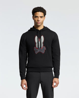 A person dressed in the MENS BULDER HOODED SWEATER - B6E261E200 from Psycho Bunny stands against a plain background. This black hoodie, adorned with a bunny emblem on the chest and crafted from soft Supima cotton, features a drawstring hood and pairs perfectly with black pants for a sleek, comfortable look.