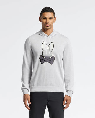 A man in a light gray Psycho Bunny MENS BULDER HOODED SWEATER (B6E261E200) with a bunny ears and crossed bones graphic stands against a plain white background, wearing black pants.