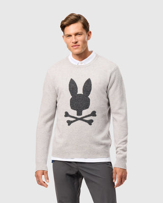 A person wearing a Psycho Bunny Men's Lincoln Merino Wool Sweater (B6E831D200) in light gray showcases a dark graphic of a rabbit head and crossbones against a plain gray background.