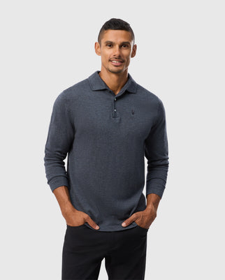 A man wearing a dark blue Psycho Bunny MENS BRADSHAW POLO SWEATER - B6E835D200 and black pants stands against a plain light gray background. He has short hair and is smiling with his hands in his pockets.