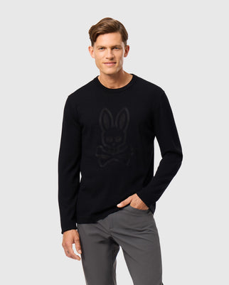 A person wearing the Psycho Bunny MENS CORY CHAINSTITCH COTTON SWEATER - B6E893D200, featuring a chainstitch embroidered rabbit and crossbones graphic on the front, stands confidently. They have short hair and pose with one hand in their pocket of gray pants against a plain light gray background.