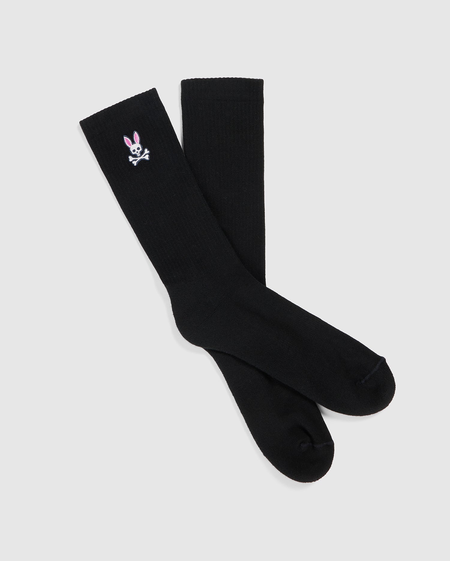 A pair of black knee-high fashion socks with a small embroidered design near the top on one sock. The design features a white skull and crossbones with pink ears resembling a bunny. Made in Peru from luxurious Pima cotton, the Psycho Bunny MENS FASHION SOCKS - B6F486C200 are displayed on a plain white background.