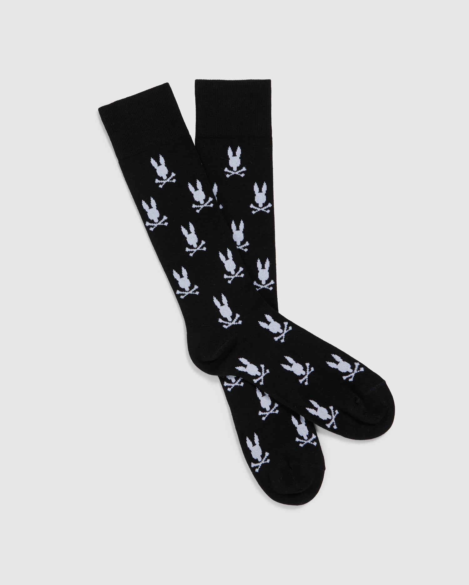 A pair of MENS DRESS SOCK - B6F750C200 from Psycho Bunny with a repeated bunny pattern in white logos throughout. Made from luxurious Peruvian Pima cotton, the socks are arranged in a crisscross pattern against a plain white background, highlighting the contrast between the black fabric and white designs.