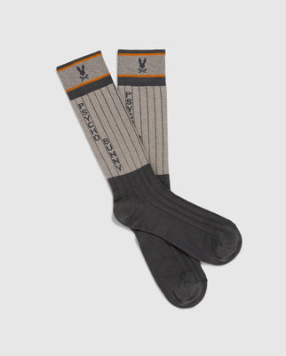 The Psycho Bunny MENS CONTRAST COLOR DRESS SOCK - B6F760D200 are knee-high dress socks in gray, featuring contrasting dark gray toes and heels with ribbed detailing. Made from soft Peruvian Pima cotton, the cuffs have an orange stripe adorned with a small bunny logo above crossed bones. The text "PSYCHO BUNNY" runs vertically down the length of the socks.