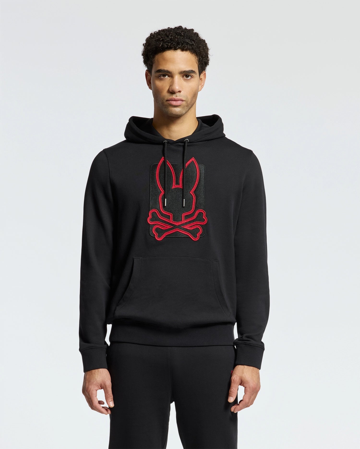 The individual is dressed in the Psycho Bunny MENS PIERCE ESSENTIAL FRENCH TERRY HOODIE - B6H135E200, a fashionable black hoodie featuring a red outline of a stylized rabbit face and crossbones design on the front. The embroidered cotton material makes it pop against the plain, light gray background.