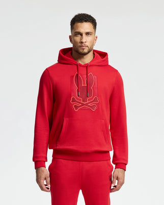 A person wearing the MENS PIERCE ESSENTIAL FRENCH TERRY HOODIE - B6H135E200 by Psycho Bunny, featuring a stylish red design adorned with a bold cartoon-style embroidery logo of rabbit ears and crossbones. This high-quality cotton hoodie is perfectly matched with red pants, all set against a plain white backdrop.