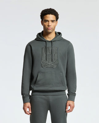 A person wearing a Psycho Bunny MENS PIERCE ESSENTIAL FRENCH TERRY HOODIE - B6H135E200, known for its stylish embroidery and high-quality cotton fabric, paired with matching sweatpants stands against a plain background. The dark grey hoodie features a stylized graphic on the chest.