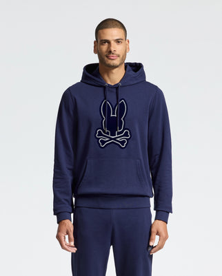 A person wearing the Psycho Bunny MENS ALEXANDER HOODIE - B6H154E200, a dark blue men's cotton blend hoodie adorned with a stylized rabbit head above crossed bones graphic. They are also sporting matching pants, making it an ideal choice for everyday wear against a plain white background.