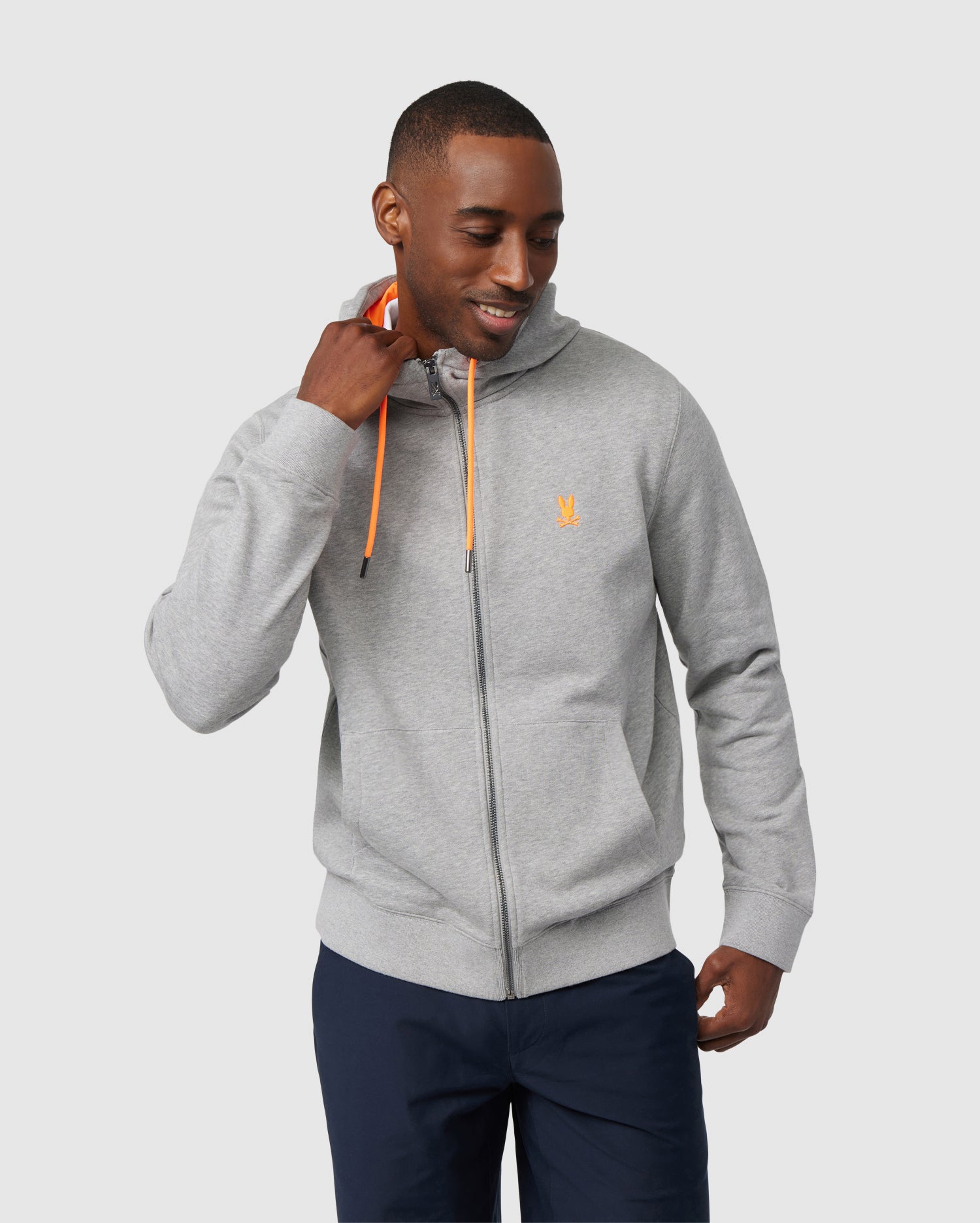 A man smiles while looking down and gently pulling on the hood string of his light gray zip-up Psycho Bunny hoodie. The MENS BUFFALO FULL ZIP HOODIE - B6H175B200 features orange drawstrings and a small orange logo on the left chest. He is also wearing dark blue pants. The background is plain light gray.