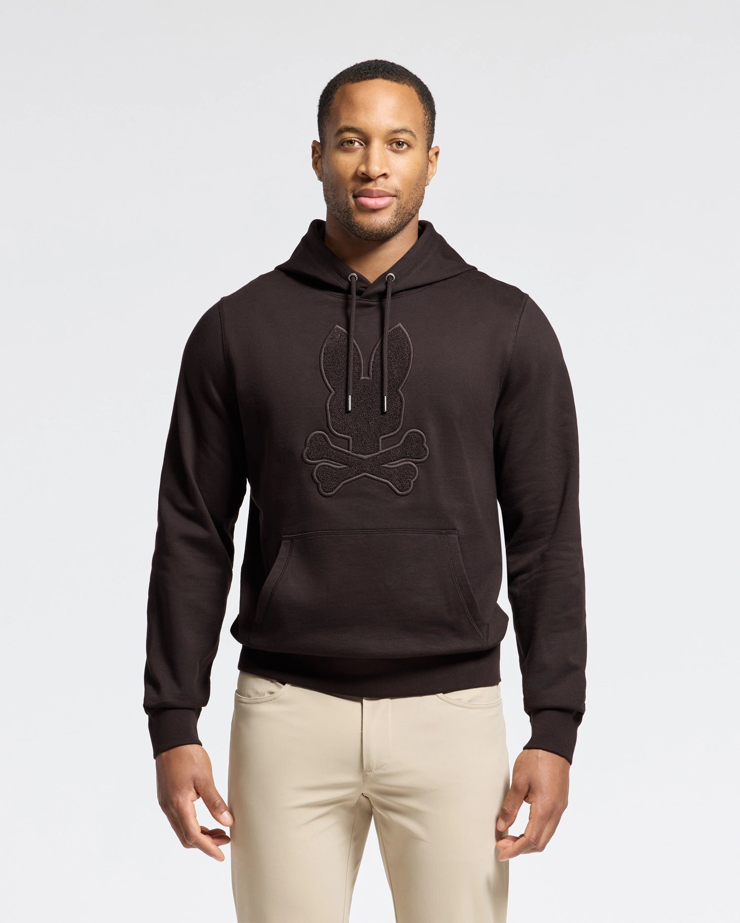 A person wearing the Psycho Bunny MENS ELDORADO BRUSH BACK HOODIE - B6H234E200, featuring a rabbit and crossbones design, stands against a plain background. Crafted from brush-back cotton with a kangaroo pocket, it complements their light-colored pants effortlessly.