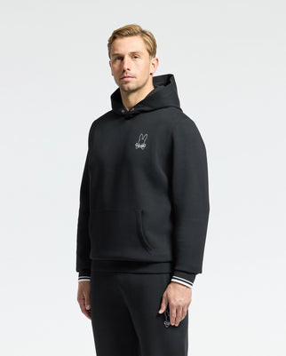 A person in a Psycho Bunny MENS DACANO PERFORATED HOODIE (B6H239E200) with a small bunny logo and striped cuffs stands relaxed. Matching black pants showcase the same bunny motif.