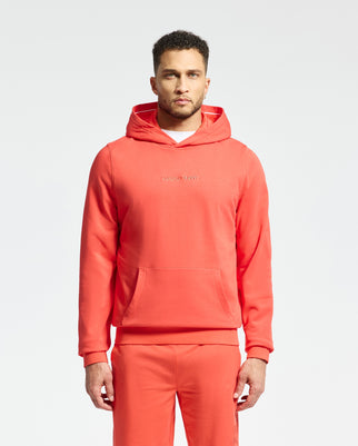 A man dons a Psycho Bunny Nora Classic French Terry Hoodie (B6H434F200) in orange, paired with matching sweatpants. The hoodie showcases an embroidered wordmark on the chest as he stands against a plain white background.