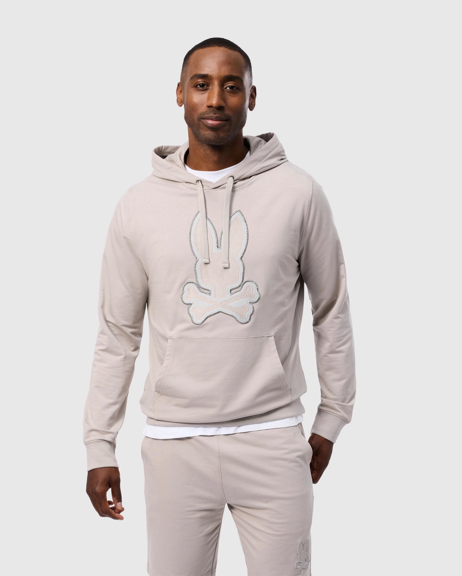 A man stands against a plain white background, wearing the MENS WALTER LIGHTWEIGHT HOODIE - B6H476D200 from Psycho Bunny. The light beige hoodie, made from Micro French terry, features a stylized bunny and crossbones graphic on the front. The regular fit hoodie has its hood pulled back, revealing his short hair. He also sports matching light beige pants and has one hand in his pocket.