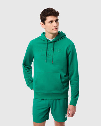 A person with short hair is wearing a matching green cotton Men's Courtside Hoodie (B6H689C200) and shorts from Psycho Bunny, featuring an embroidered logo graphic on the left leg. The regular fit hoodie includes a front pocket and drawstrings. The individual is standing against a plain light gray background.