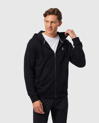 A person wearing a black Psycho Bunny MENS LEVY ZIP FLEECE HOODIE - B6H720D200 over a white t-shirt stands against a plain background. They are slightly smiling and have one hand adjusting the hoodie's drawstrings. The hoodie features a small logo on the left chest.