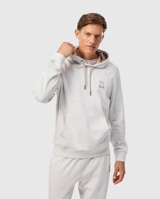 A person wearing the MENS JAMES PREMIUM HOODIE - B6H789D200 by Psycho Bunny in light gray, paired with matching sweatpants, stands against a plain background. Crafted from premium jersey for stylish warmth, the hoodie features a small embroidered bunny design on the left side. The person has short hair and is resting one hand near the hood.