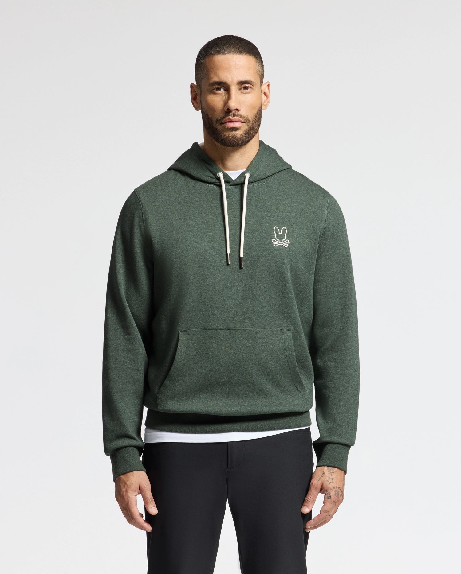 A man with a beard is stylishly dressed in a trendy dark green MENS JAMES PREMIUM HOODIE - B6H789D200 from Psycho Bunny, featuring a small logo on the chest. Paired with black pants, this high-quality men's cotton hoodie enhances his style as he stands against a plain white background, looking directly at the camera.