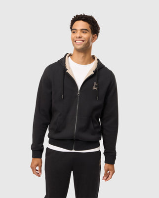 A smiling man wears a Psycho Bunny MENS JAMES PREMIUM ZIP HOODIE - B6H793D200 over a white t-shirt and matching black sweatpants. The hoodie features an embroidered Bunny graphic on the left chest area. He appears to be standing against a plain, light-colored background.