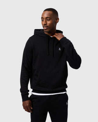 A person wearing a black Psycho Bunny MENS LEVY FLEECE HOODIE - B6H859D200 and black pants stands against a plain white background. The 100% cotton hoodie features a small white logo on the left chest. The individual is smiling slightly and holding the drawstring of the hoodie with one hand.