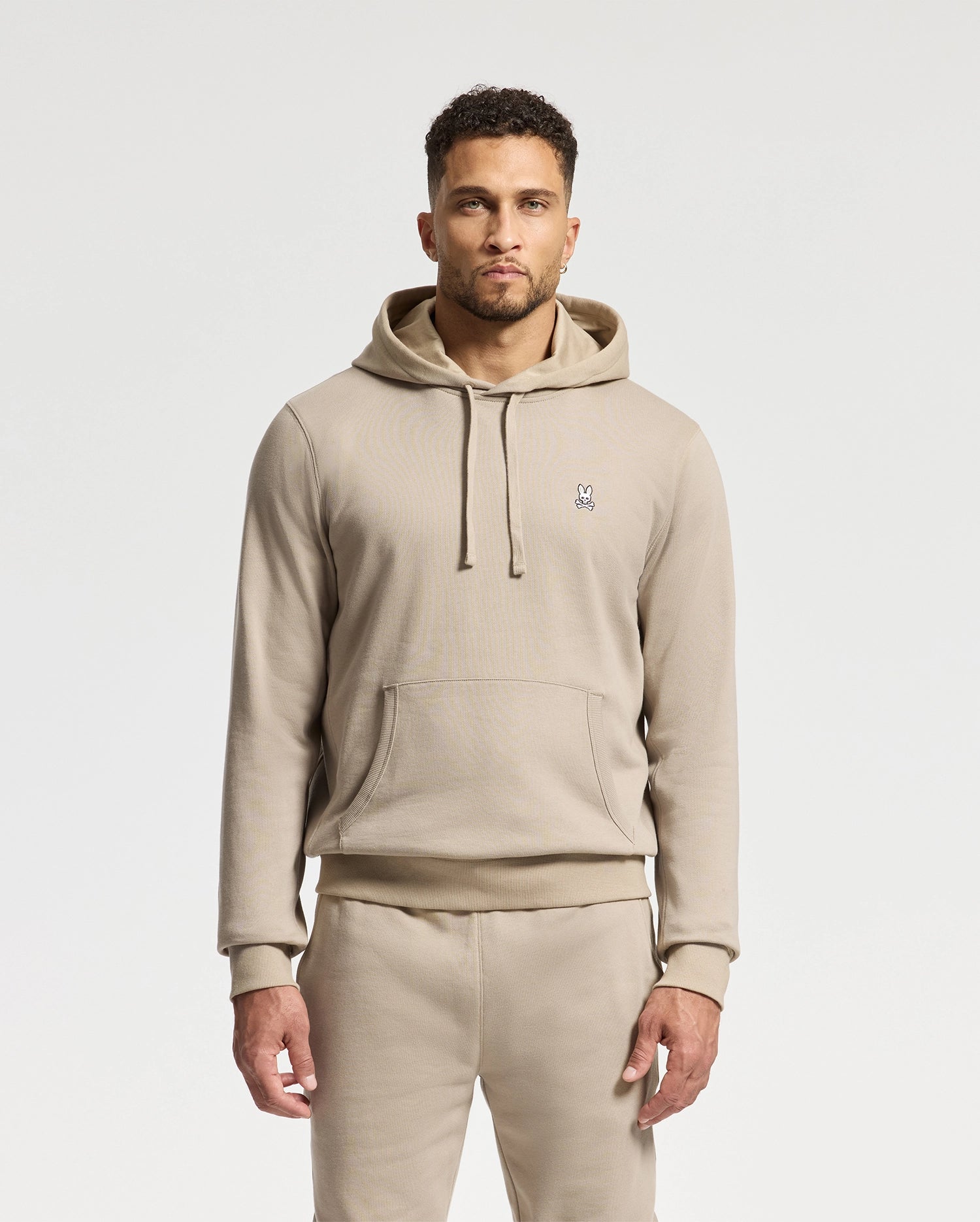 A man sporting a Psycho Bunny MENS LEVY ESSENTIAL FLEECE HOODIE in beige, paired with matching sweatpants, stands against a plain background. The hoodie features a small embroidered rabbit logo on the chest. He has short, curly hair and a neatly trimmed beard.