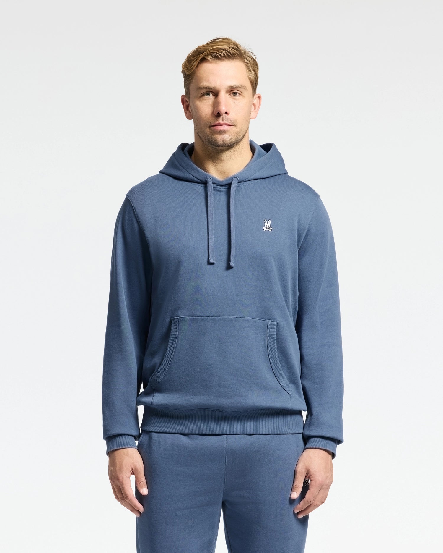 A person in the Psycho Bunny Men's Levy Essential Fleece Hoodie and matching sweatpants stands facing forward against a plain white background. The blue hoodie, crafted from 100% cotton French terry, features a small emblem on the chest. The person has short blond hair.