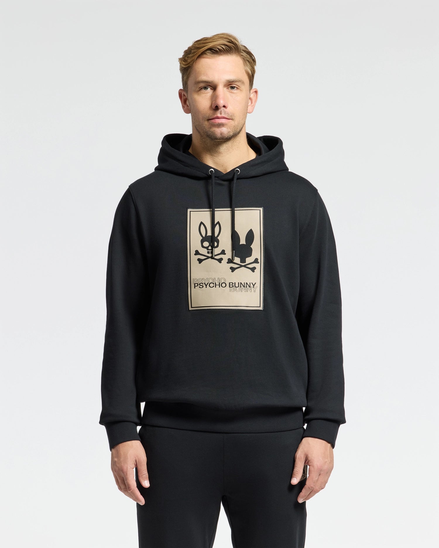 A man with short blond hair stands in front of a plain white background, exuding comfort and ease in his stylish black MENS MONROE TWILL PATCH FLEECE HOODIE - B6H926D200 from Psycho Bunny, which features two rabbits and the brand name on the front.