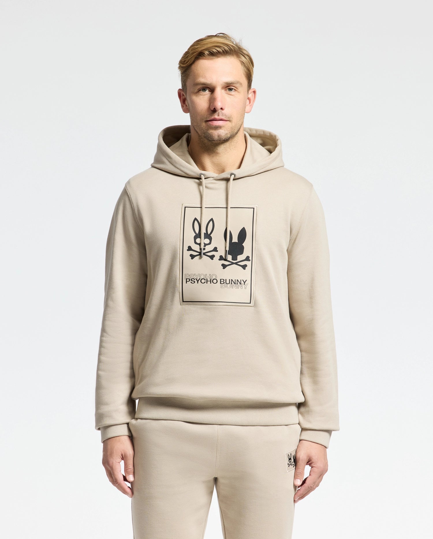 A man wearing the MENS MONROE TWILL PATCH FLEECE HOODIE from Psycho Bunny, along with matching sweatpants, is standing against a plain background. The outfit features the 