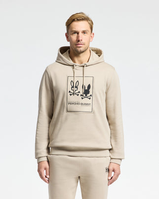 A man wearing the MENS MONROE TWILL PATCH FLEECE HOODIE from Psycho Bunny, along with matching sweatpants, is standing against a plain background. The outfit features the "Psycho Bunny" logo appliqué. He has short blond hair and is looking directly at the camera.