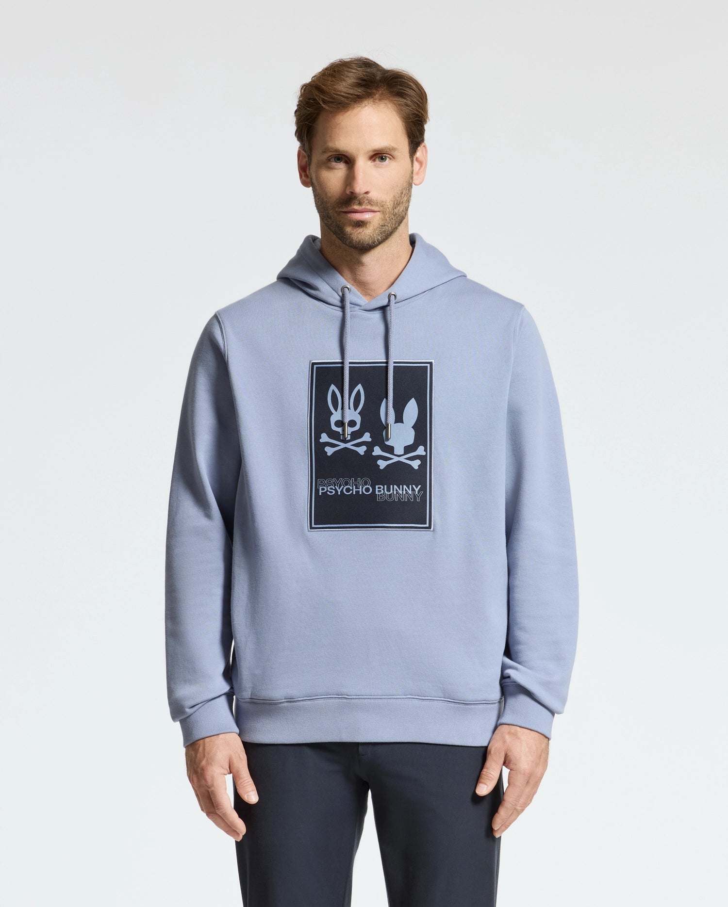 A man wearing a light blue Psycho Bunny MENS MONROE TWILL PATCH FLEECE HOODIE - B6H926D200, showcasing the iconic bunny and crossed bones logo appliqué. The French terry fabric pairs perfectly with his short hair and beard as he stands against a plain white background.