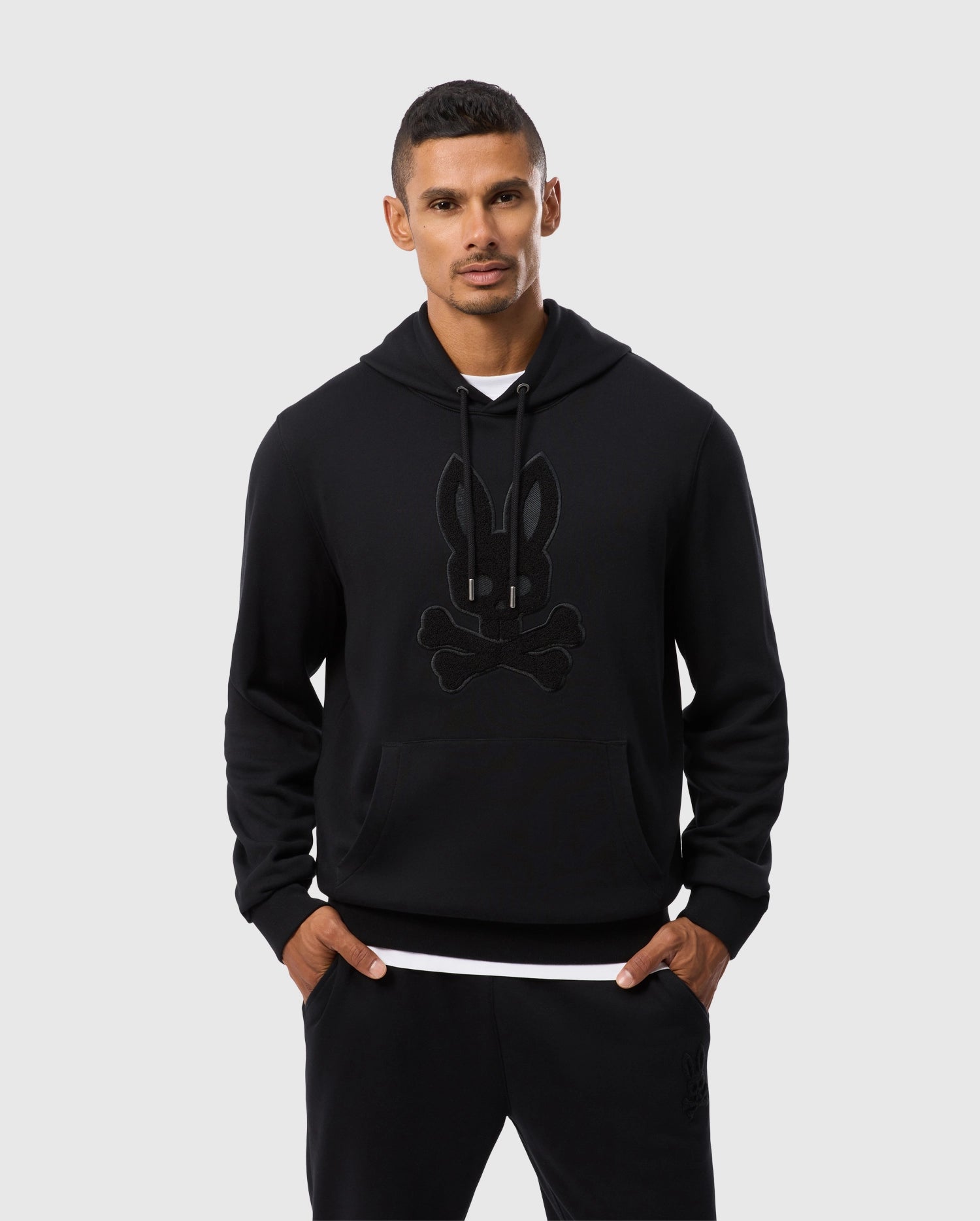 Wearing the Psycho Bunny MENS PACIFIC CHENILLE HOODIE (B6H930D200), featuring a stylized rabbit and crossbones logo, a man stands with his hands in his pockets. The chenille embroidery adds texture to the design as he poses against a plain white background.