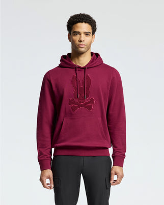 A man stands against a plain light background, wearing the luxurious Psycho Bunny MENS PACIFIC HIGH PILE CHENILLE HOODIE - B6H930D200 in maroon, featuring a black rabbit and crossbones design. His curly hair frames his face as he looks directly at the camera.