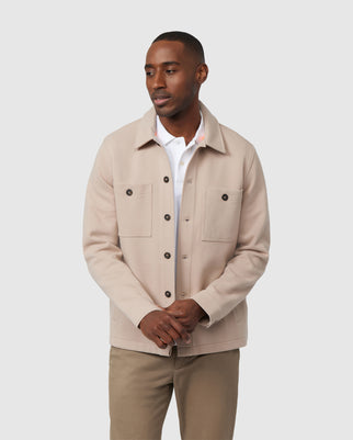 A man wearing a light beige Psycho Bunny MENS BEDFORD SHIRT JACKET - B6J290B200 over a white shirt stands against a plain light background. The jacket showcases logo-engraved buttons and work-jacket style pockets, and he gazes to his left with a relaxed expression.