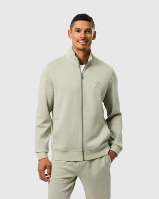 A person in a light green track-style outfit, consisting of the Psycho Bunny MENS LYONS HIGH COLLAR FULL ZIP (B6J380B200) and matching pants, stands smiling with hands in pockets. The full-zip sweatshirt showcases a subtle stitched design on the chest, adding unique flair against the plain white background.