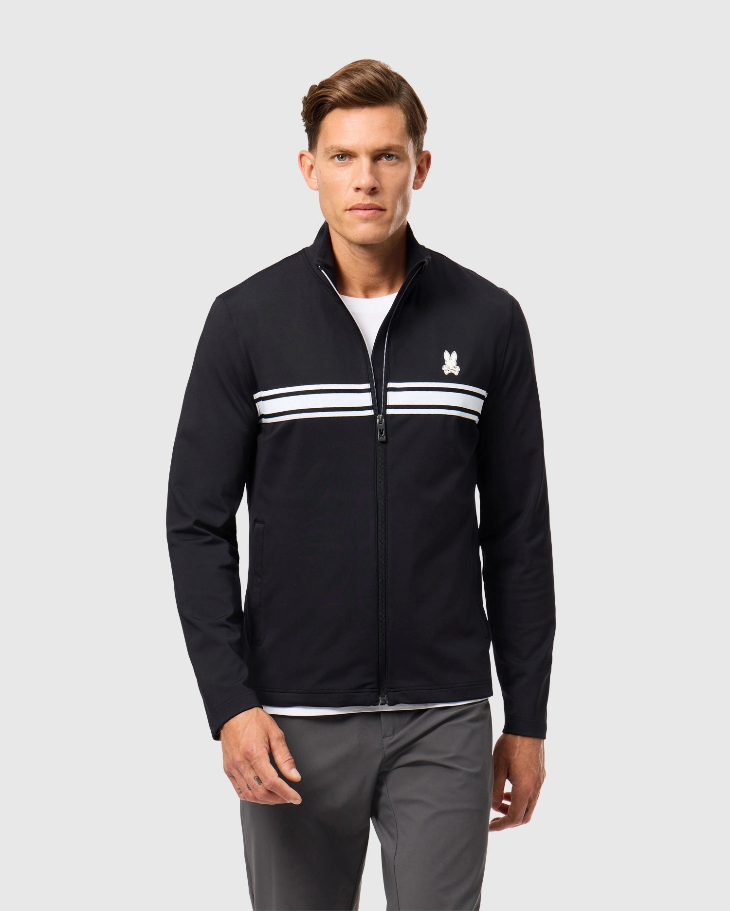 A man is wearing the Psycho Bunny MENS TERRELL SPORT ZIP UP JACKET - B6J643C200, showcasing its versatile design with white horizontal stripes across the chest and a small logo on the left side. He pairs it with a white shirt and gray pants, standing against a plain gray background to highlight the jacket's sleek style.