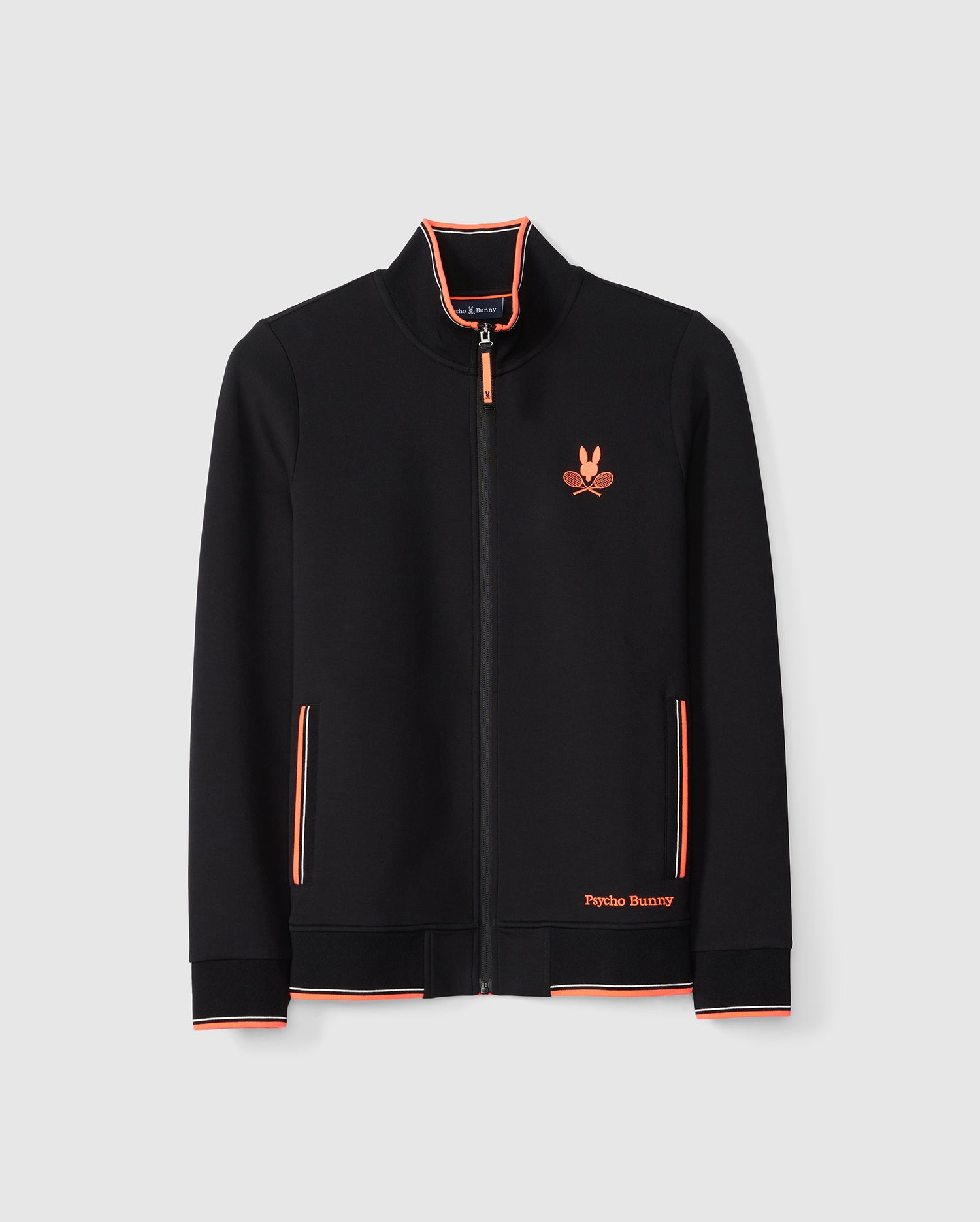 A black MENS COURTSIDE TRACK JACKET - B6J692C200 by Psycho Bunny is displayed against a white background. The jacket features orange and white striped accents on the collar, cuffs, and pockets. An embroidered orange logo with a bunny and crossed racquets graces the chest, accompanied by 