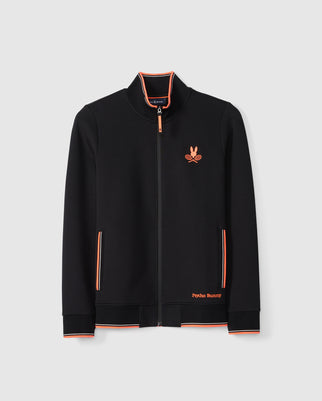 A black MENS COURTSIDE TRACK JACKET - B6J692C200 by Psycho Bunny is displayed against a white background. The jacket features orange and white striped accents on the collar, cuffs, and pockets. An embroidered orange logo with a bunny and crossed racquets graces the chest, accompanied by "Psycho Bunny" text near the hem.