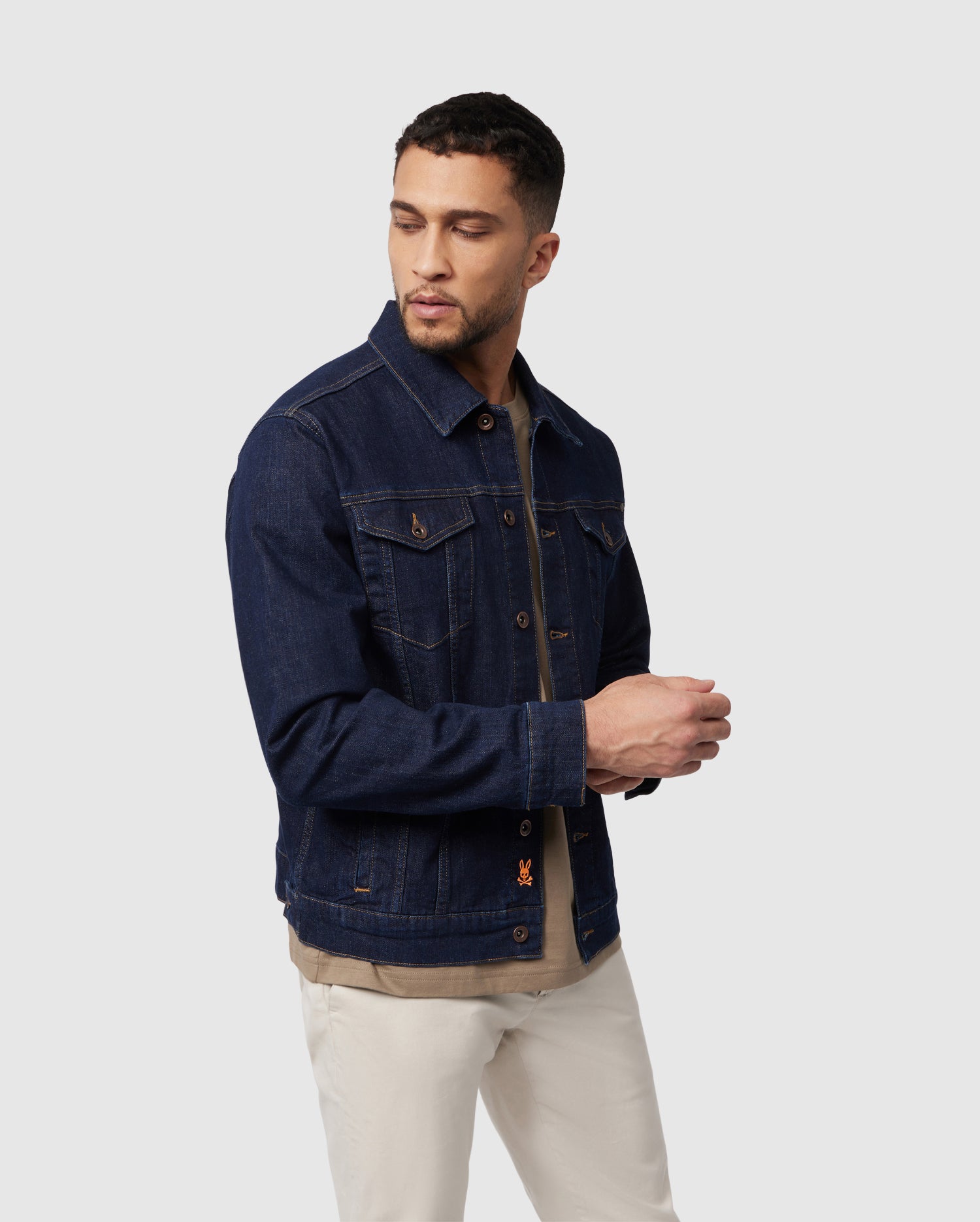 A man styled in the Psycho Bunny MENS PATHE DENIM JACKET - B6J741X1CO, layered over a beige shirt and paired with white pants, stands against a light gray background. His tranquil expression and extended hand exude modern men's fashion with understated elegance.