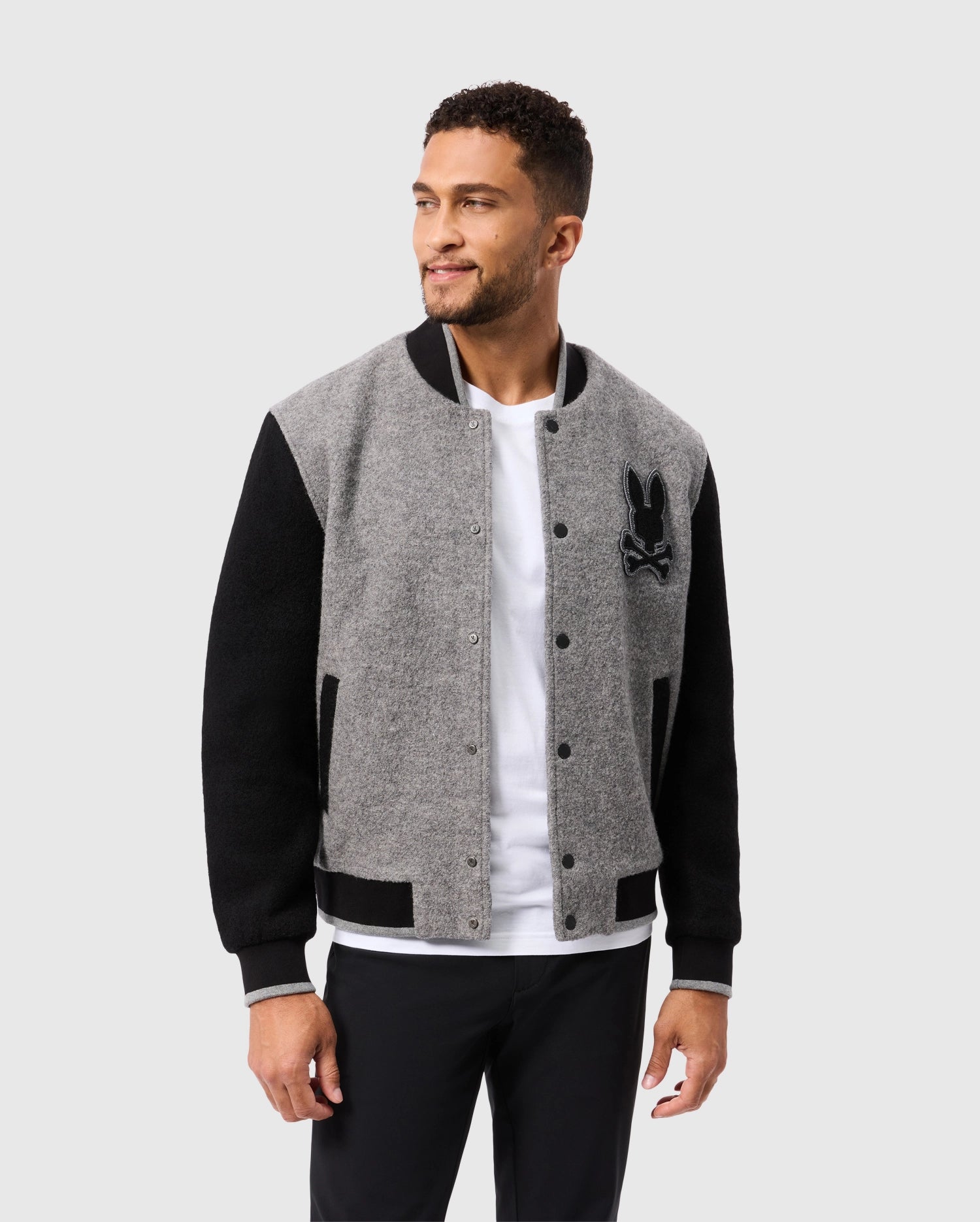 A man stands elegantly in a Psycho Bunny MENS TOM WOOL BOMBER JACKET - B6J744D200, adorned with a rabbit logo, casting a sideways glance. His short hair and beard add to the sophistication of his grey and black outfit, which includes black pants and a white t-shirt. The subtle light grey background accentuates his polished appearance.