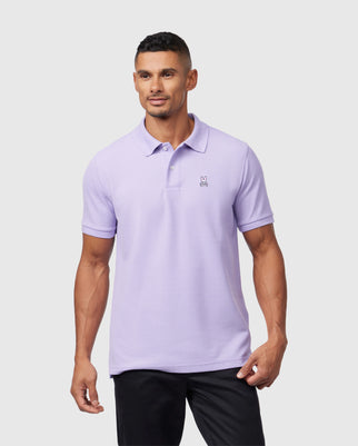 A man wearing a light purple Psycho Bunny pima cotton polo shirt with a small logo on the left chest, standing against a plain white background. He has a short haircut and a fit build.