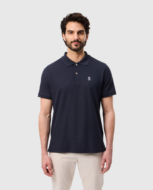 A man with dark hair and a beard is wearing the Psycho Bunny Men's Classic Pique Polo Shirt (B6K001ARPC) in dark navy, made from 100% Pima cotton, featuring a small embroidered logo on the left chest. He is standing and facing the camera with a neutral expression, sporting beige pants against a plain white background.