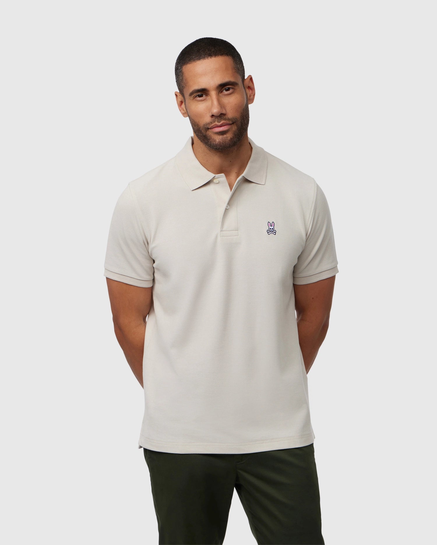 A man with short hair and a beard is wearing a luxurious Psycho Bunny MENS CLASSIC PIQUE POLO SHIRT - B6K001B200 with a small embroidered rabbit logo on the chest. Standing against a plain white background with his hands behind his back, he completes his timeless look with dark green pants.