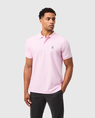 A man stands against a plain grey backdrop, wearing the Psycho Bunny MENS CLASSIC PIQUE POLO SHIRT - B6K001CRPC in a light pink hue, made from Peruvian Pima cotton, featuring a small black bunny emblem on the left chest. He has short curly hair and is also wearing dark grey regular-fit pants, looking slightly off to his left.