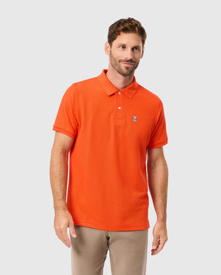 A man with short hair is wearing the MENS CLASSIC PIQUE POLO SHIRT - B6K001CRPC by Psycho Bunny, in orange, paired with beige pants, as he stands against a plain gray background.