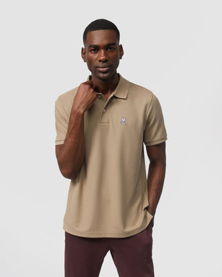 A man in a beige Psycho Bunny Pima cotton polo shirt and dark pants stands against a white background, lightly tugging on the collar of his shirt, and looking towards the camera.